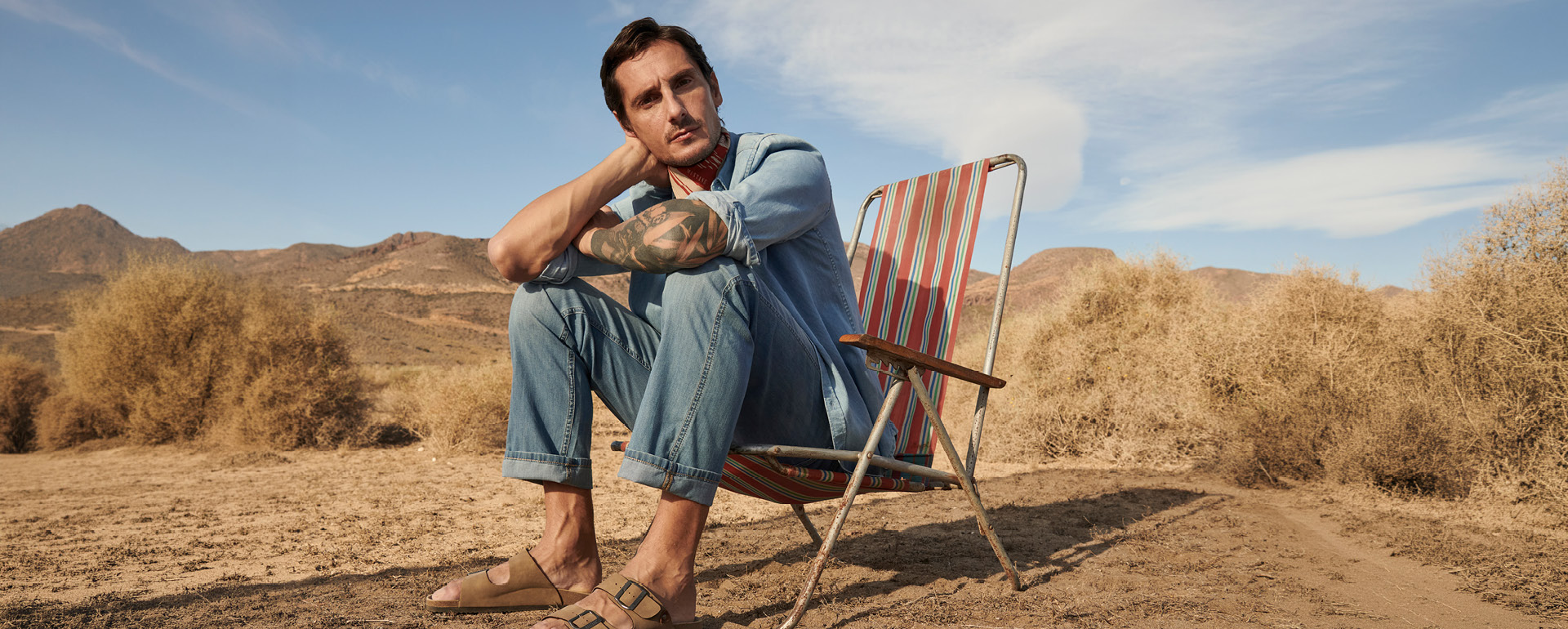 denim shirt, denim jeans, tanned sandals, desert chair, tattooed arm